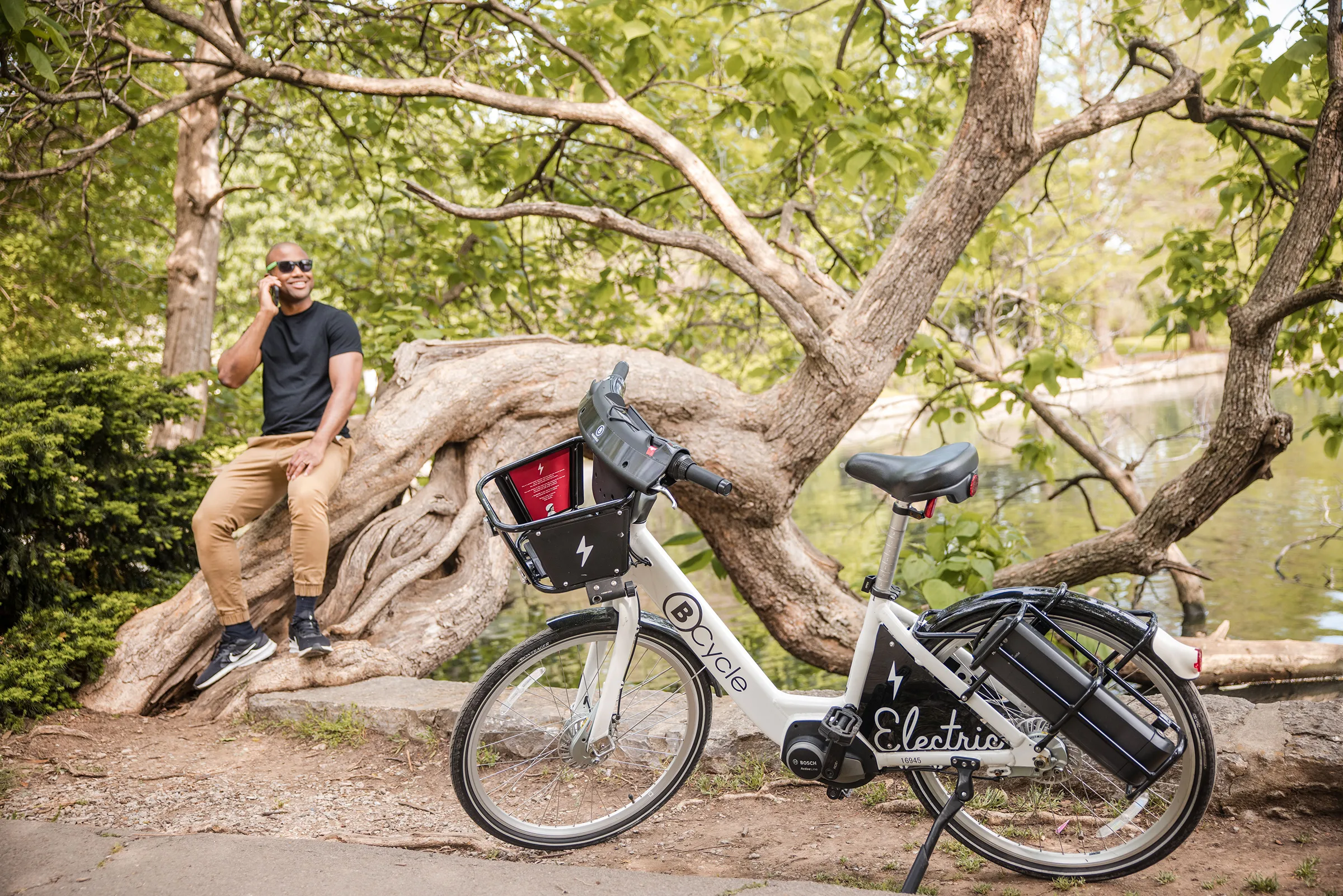 Photo of man sitting on a tree by a pond next to his bike share bicycle