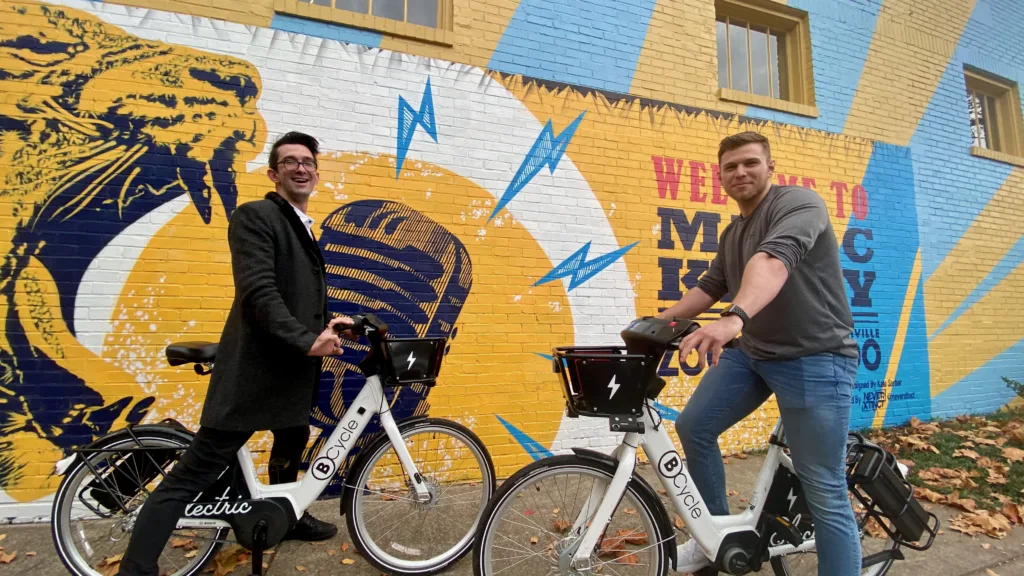 Photo of two men riding bike share bicycles in front of a music city mural