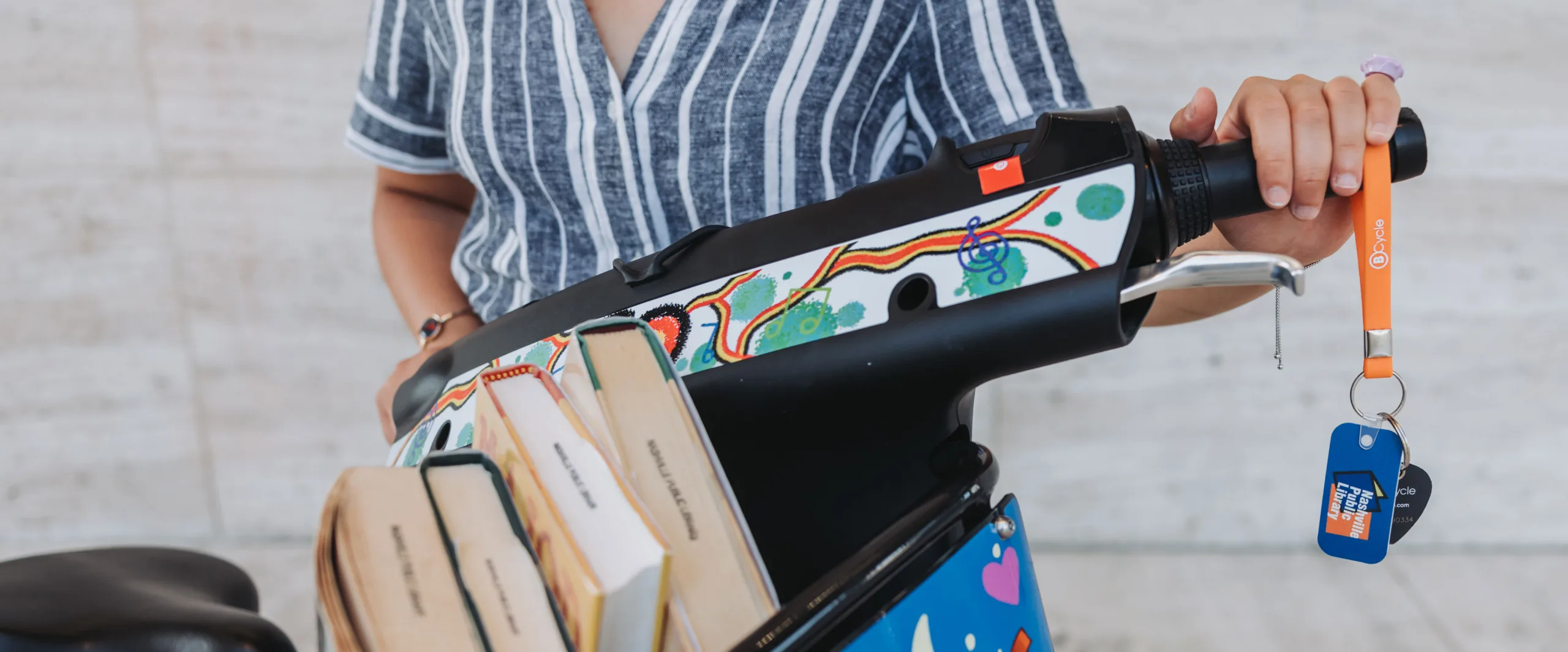 girl riding a bike share bicycle with a basket full of library books while holding her Nashville Library card